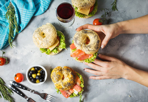 Bagels with salmon, vegetables, cream-cheese and glass of red wine on grey concrete background. Woman hands hold bagel