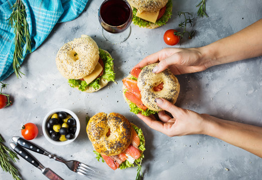 Bagels with salmon, vegetables, cream-cheese and glass of red wine on grey concrete background. Woman hands hold bagel