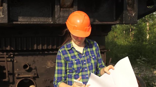 A young woman worker in orange helmet stands near a mining excavator, looks in the project then talking on the phone.