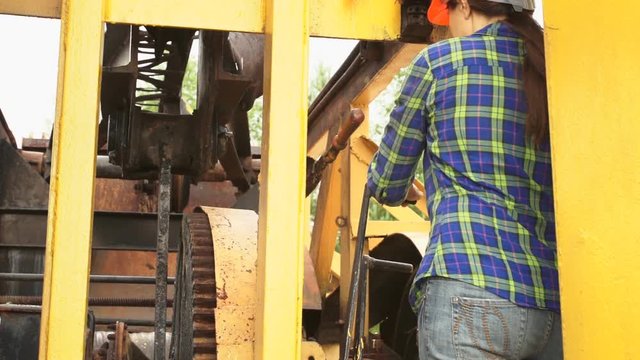 A young woman worker in orange helmet checks the mechanisms of the old yellow excavator warm day.