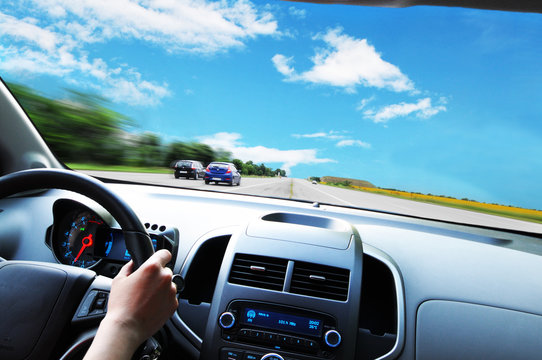 Car dashboard with driver's hand on the steering wheel against road and sky with clouds