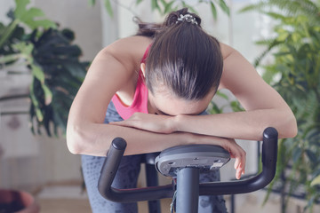 Young healthy fit woman training at home on exercise bike during work-out feeling exhausted and...