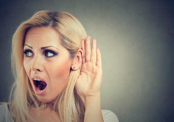 Shocked woman holds her hand near ear and listens carefully isolated on gray background 
