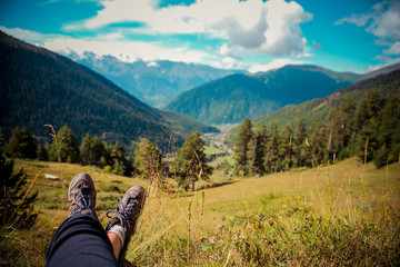 Fototapeta na wymiar Hiker rest and chilling in the grass to beautiful mountain view. Legs with hiking boots. Caucasus Mountains. Svaneti, Geogria.