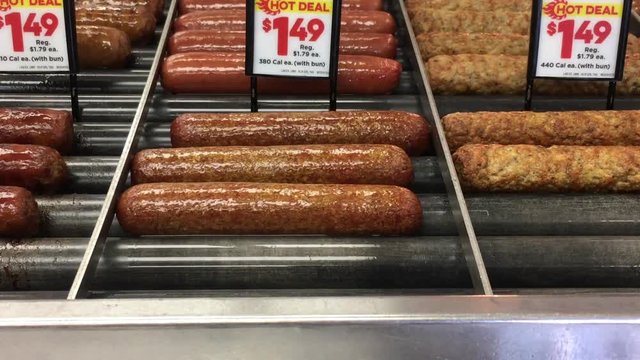 Sausages and hot dogs are spinning on a roller machine and displayed under bright retail lighting with deal pricing. 