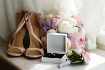 Wedding rings in white box, beige sandals, a bouquet of flowers and boutonniere at the window. Accessories for wedding. The symbols of love, wedding rings,shoes, wedding bouquet with boutonniere