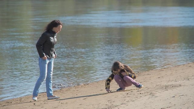 A happy family walks by the river. A little girl draws on the sand near the river.