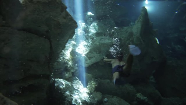 underwater landscape, man in swimsuit with fins dive from camera in stream of sun and make many bybbles, group of snorkeling people swiming with flashlight on the background in cenote cave dos ojos 