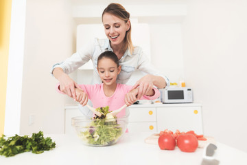 Mom and daughter have fun while preparing a salad. They are in a bright kitchen.