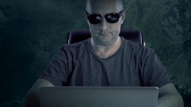 A young man in a T-shirt and glasses is sitting at the computer. A man works on a computer late in the dark at night and coughs. The manager works a lot.