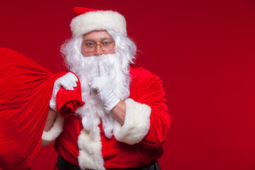 Portrait of Santa Claus with huge red sack keeping forefinger by his mouth and looking at camera