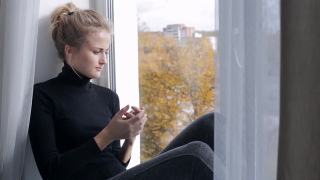 Sad young woman with smartphone sitting on windowsill