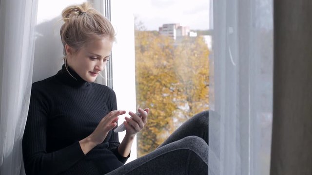 Young woman using her smart-phone sitting on windowsill