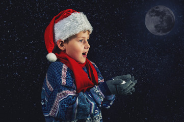 Fototapeta na wymiar Happy boy in Santa hat plays with snowflakes on a dark background . Happy Christmas holidays