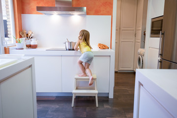 Four years old blonde child climbing on stool o ladder to cook in electrical cooktop with a...