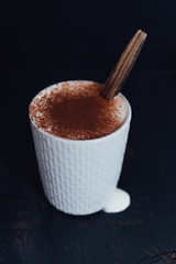 Light grey stoneware mug with stiched pattern filled with hot chocolate with slightly whipped cream and dusted with cocoa powder on black background and wooden spoon