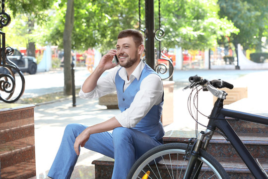 Handsome businessman with bicycle sitting on stairs and talking by mobile phone outdoors