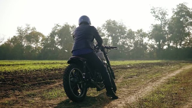 Back view of a man in helmet and leather jacket coming up to his bike and starting the engine while standing on the offroad on a sunny day. Slowmotion shot