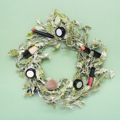 Christmas wreath with makeup on green background, flat lay with lipstick, eyeshadows, nail varnish