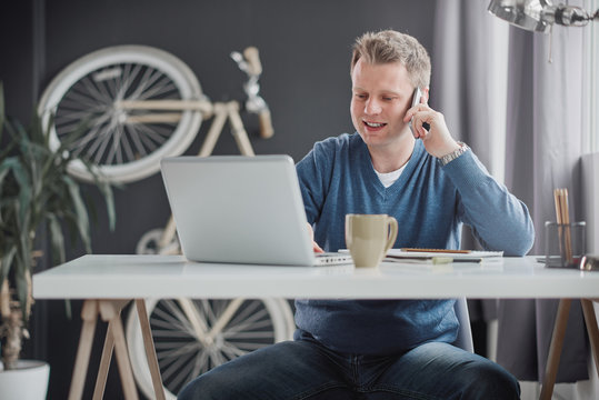 Businessman holding in one hand smart phone and with other typing on keyboard. He is siting at modern home office