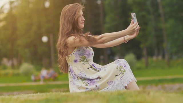 Beautiful young girl in summer dress sitting on grass in park, takes photo of herself using smartphone. The girl has beautiful long hair. Summer. Recreation.