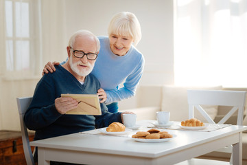 Show it to me. Relaxed old man sitting in the dining room and drinking coffee while looking at little picture