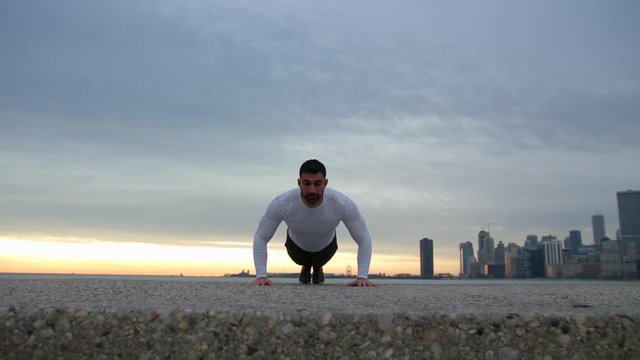 Man is doing push ups on the ebankment. Athlete training outdoors. Downtown with skyscrapers on the background. Seaside sunrise.