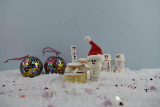 christmas food photography image using marshmallows shaped as snowman with happy iced on smiles standing in snow with sponge cream fairy cake and bauble tree decorations in the background