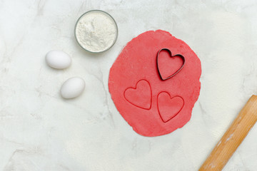 Red dough with a cut out hearts, eggs, flour and rolling pin,on a white table. Top view