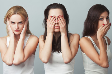 Close-up portrait of three young women in white sleeveless shirts imitating see no evil, hear no evil, speak no evil concept on gray background. Human emotions, expressions, communication. Studio shot