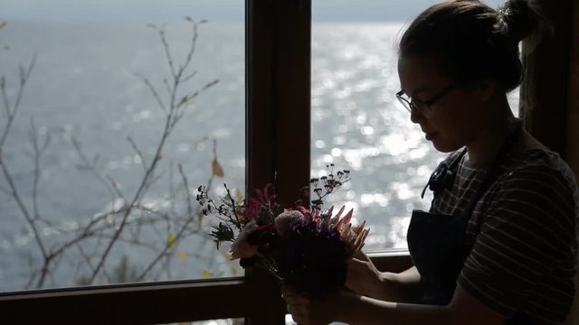 Young woman makes floral bouquet standing by window. Talented florist composes flower composition, inserting fresh blossoms and smiling amicably. Female in glasses is engaged in favorite business with