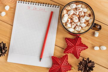 Cup of hot cocoa or chocolate with marshmallow, holiday decorations red stars and notebook with to do list on wooden table, christmas planning concept.