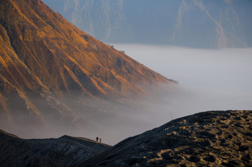 Two persons seen from behind walk along the edge of a volcano crater in famous geologic formation Gunung Bromo, Java, Indonesia.