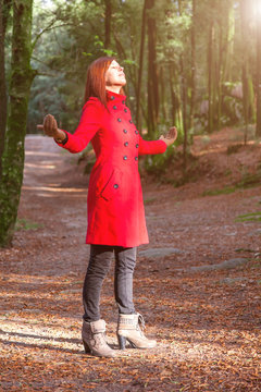Woman enjoying the warmth of winter sunlight alone on forest park path with arms open receiving rays of light, wearing a red long coat or overcoat