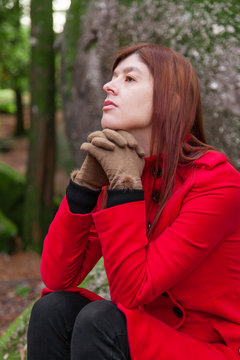 Depressed and sad young woman feeling depression sitting on forest, looking up with melancholic thinking, wearing a red long coat or overcoat during fall, autumn or winter