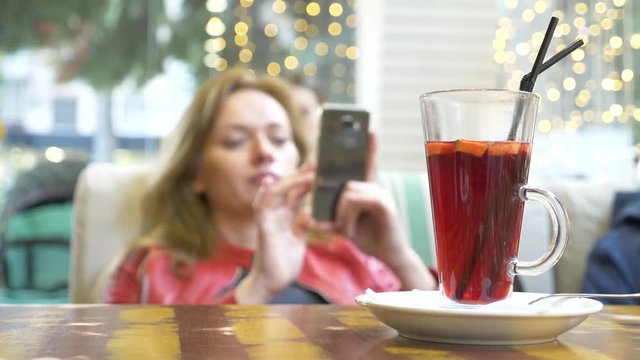 beautiful woman is resting in a cafe, drinking hot mulled wine and looking into her smartphone. 4k, background blur.