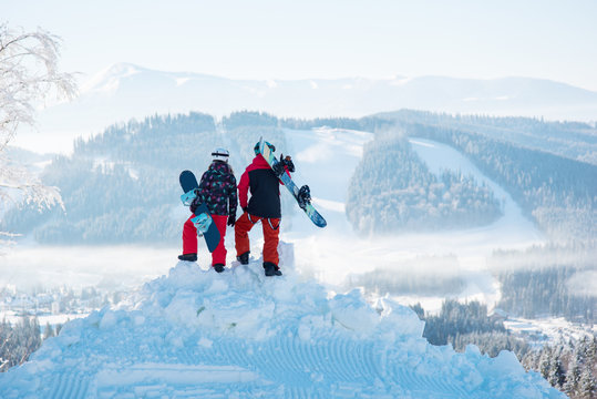 Couple of snowboarders enjoy the snow-white scenery of mountains and forests of the Carpathians from the height of the mountain top at winter ski resort on a beautiful sunny day. Back view