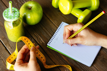 Woman writing nutrition diet and fitness workout routine. Healthy green detox, apple and dumbbells for slimming down concept. Female hands with measuring tape for checking weight loss.