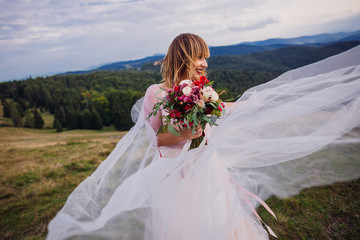 Fototapeta na wymiar Bride in white dress whirls before beautiful mountain landscape holding red wedding bouquet