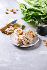 Ingredients for cooking classic Caesar salad. Sliced baked chicken breast, green roman salad, parmesan cheese, boiled egg, croutons, salt, jug of sauce over gray texture background. Copy space.