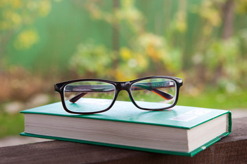 Closed green book, glasses on a table in an autumn garden. Yellow leaves.