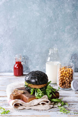 Homemade burger in black buns with avocado, arugula, cheese, onion on wood serving board with bottle of ketchup, milk, roast chickpeas over white wooden plank table. Homemade fast food.