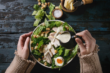 Female hands powring dressing to Classic Caesar salad with chicken breast in white ceramic plate....