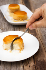Woman using spoon cut homemade caramel custard cake on white plate. Sweet dessert. Served on wooden table.