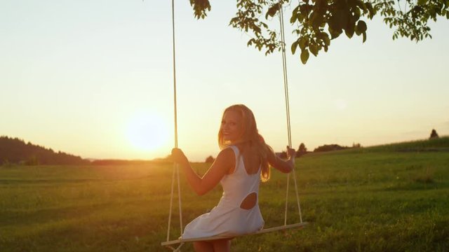 SLOW MOTION LENS FLARE: Smiling young woman swaying on rope swing under big tree at sunrise. Pretty girl in white dress swinging over the sun at golden sunset. Beautiful lady enjoying summer evening