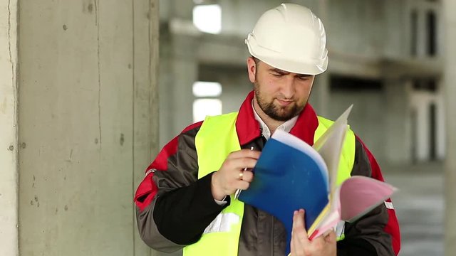 Building inspector with construction documentation at project site. Master builder in white hard hat with works papers at construction site