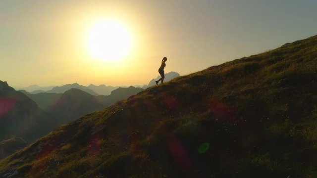AERIAL SILHOUETTE: Camera flying along a silhouette of woman hiker walking up a grassy hill in front of gorgeous yellowish landscape. Young female ascending a mountain peak on sunny summer evening.