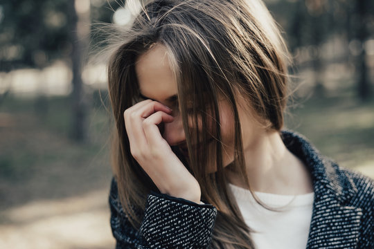 Pretty, young and shy woman covers her face with her hand. Model posing on camera in public park. Girl dressed in grey topoat. Outdoor.