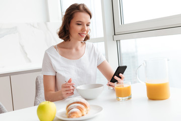 Obraz na płótnie Canvas Young attractive woman in white tshirt using mobile phone while having breakfast at the kitchen