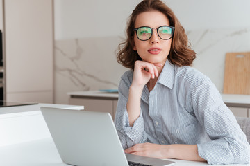 Close-up photo of beautiful brunette girl in glasses holding her chin and looking aside while sitting at workplace